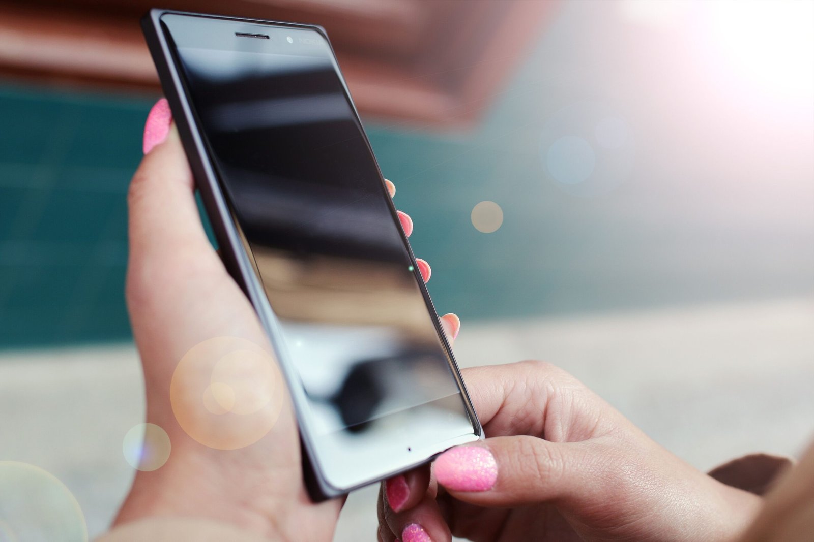 Close-up of hands holding a smartphone outdoors with a bright lens flare and reflections.
